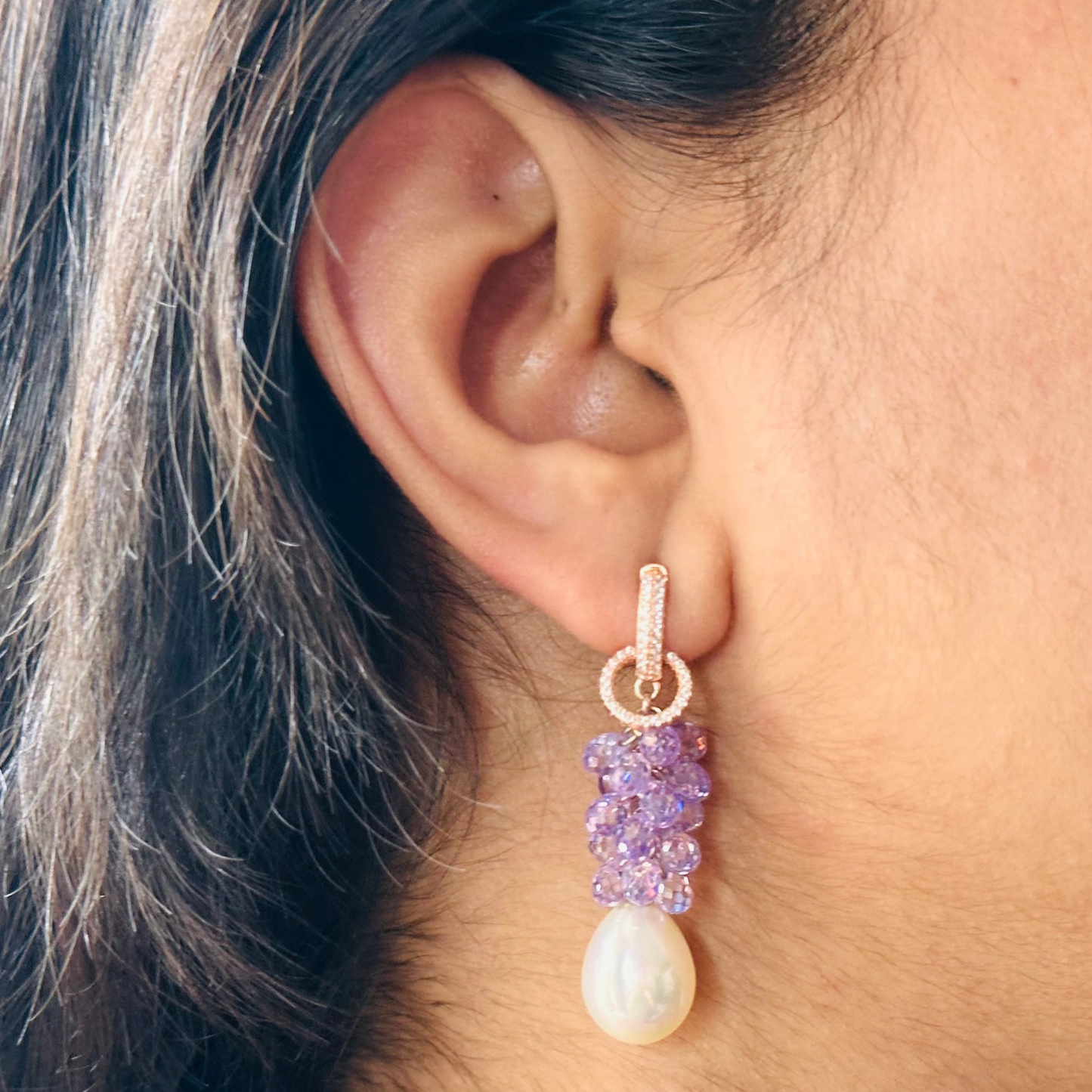 A close-up image of a woman's ear wearing a pearl drop earring with amethyst gemstones and a rose gold-plated hoop.