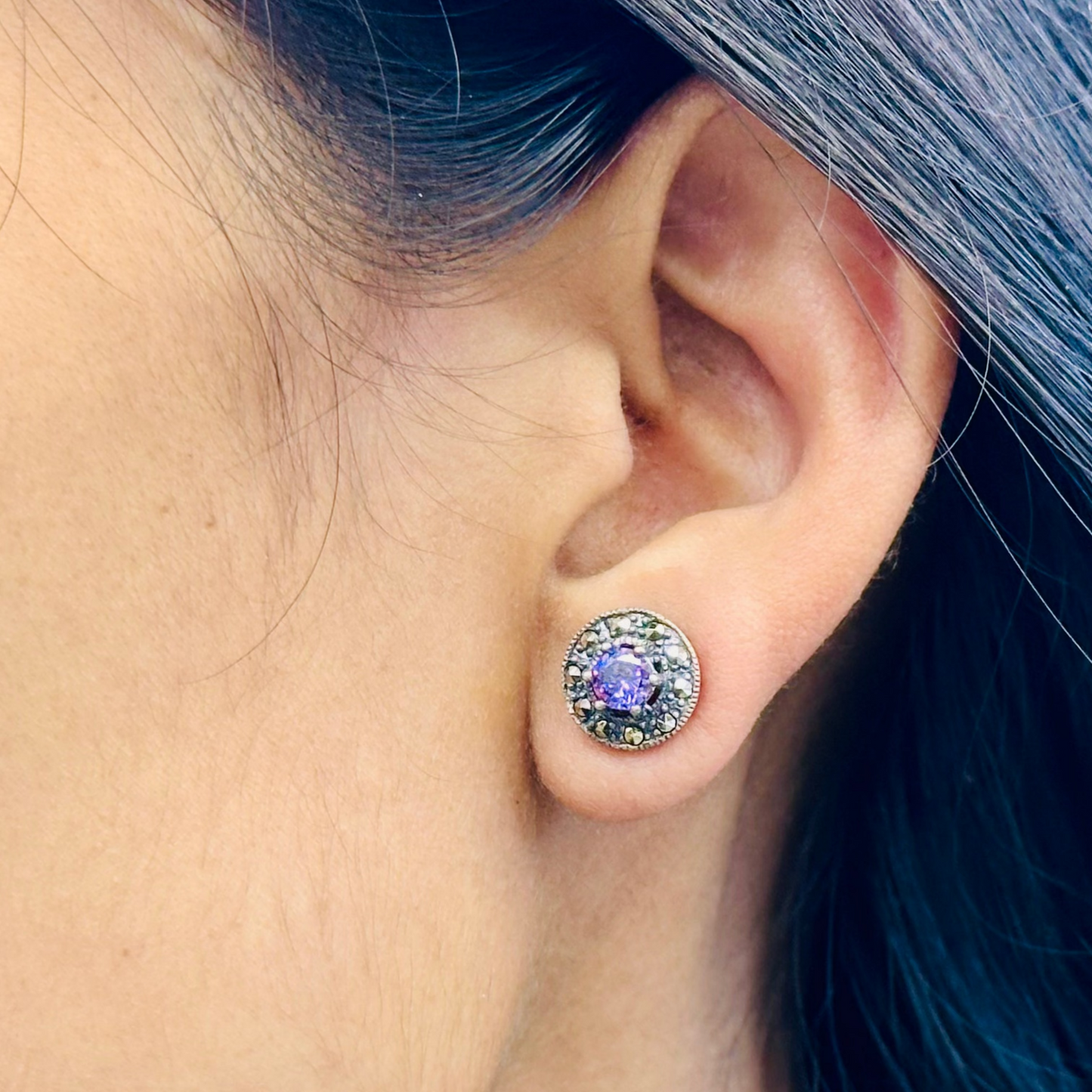 Close-up of an ear wearing a stud earring with a purple gemstone, hair color is blue.