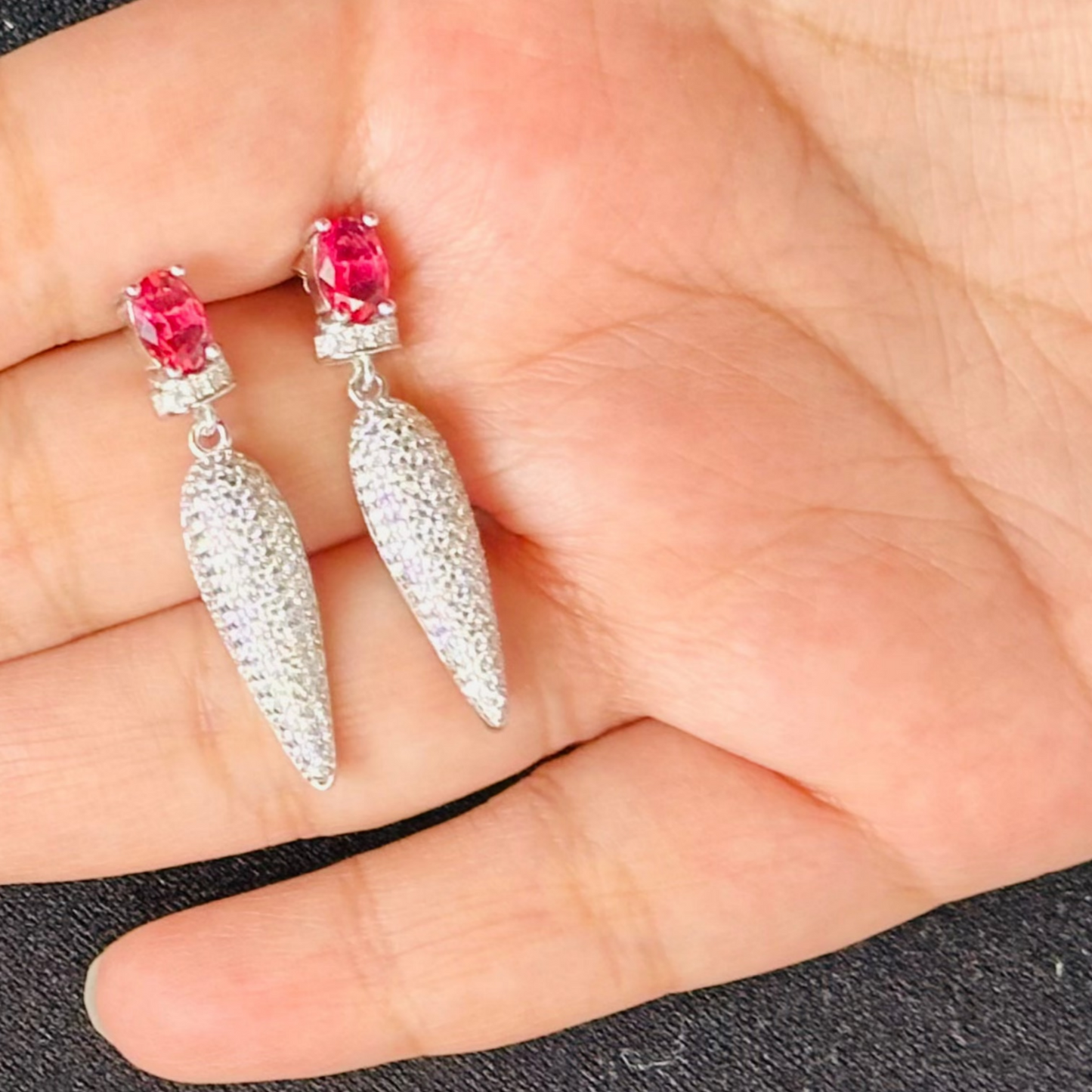 a pair of ruby red, drop-style earrings made of sterling silver. The earrings feature a central ruby gemstone with a micro-paved cubic zirconia detail.