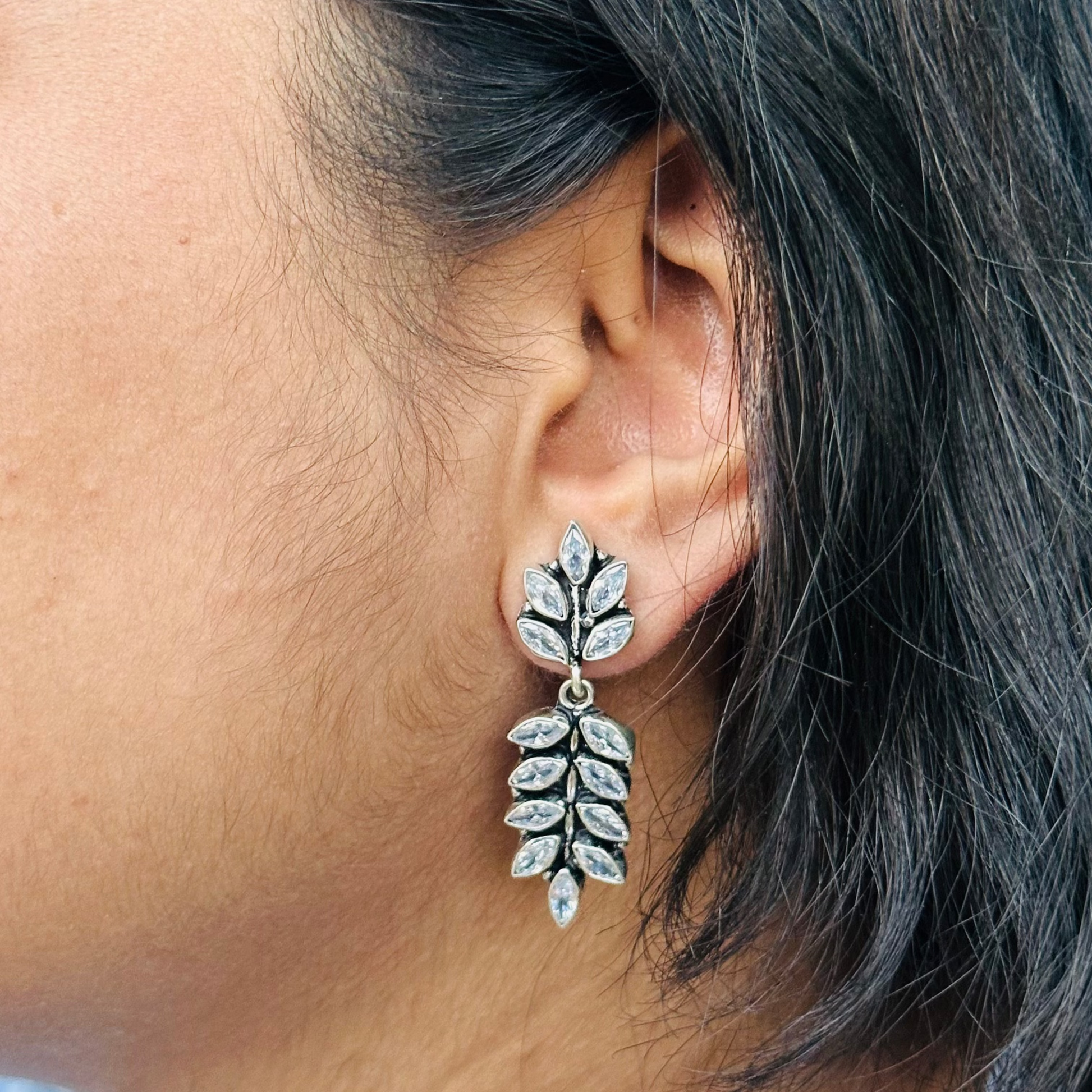 A close-up image of a woman's ear wearing a silver leaf-shaped earring adorned with white cut stones.