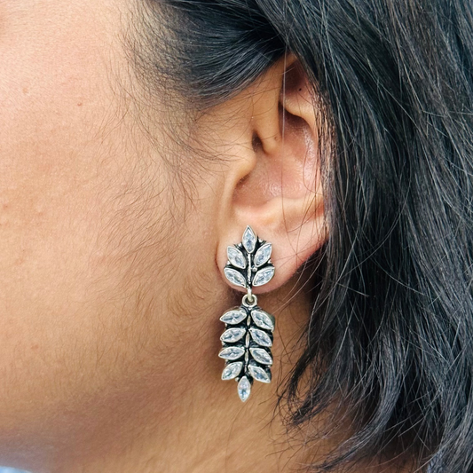 A close-up image of a woman's ear wearing a silver leaf-shaped earring adorned with white cut stones.