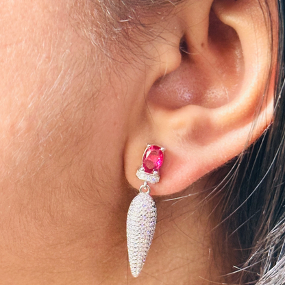 Showcasing elegant ruby drop earrings, the image features a pair of sterling silver earrings. A vibrant ruby gemstone is the focal point, complemented by a sparkling cascade of micro-paved cubic zirconia, creating a luxurious and eye-catching design.