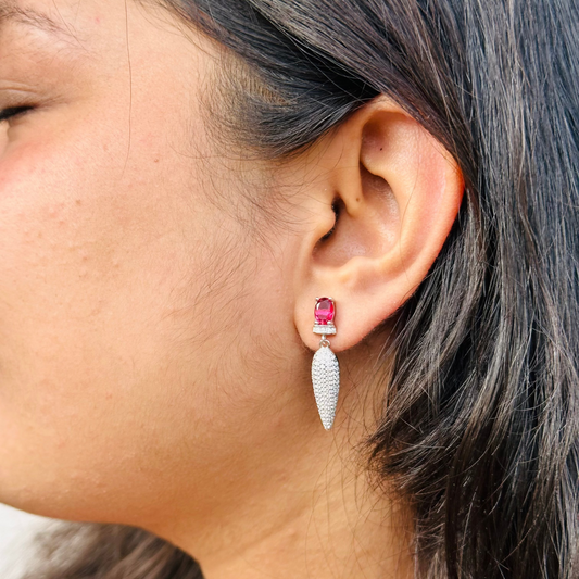 The image showcases a pair of elegant, drop-style earrings. They feature a prominent, ruby-red gemstone as the focal point, complemented by a cascade of sparkling, micro-paved cubic zirconia, all set in a silver base. The overall design exudes sophistication and luxury.